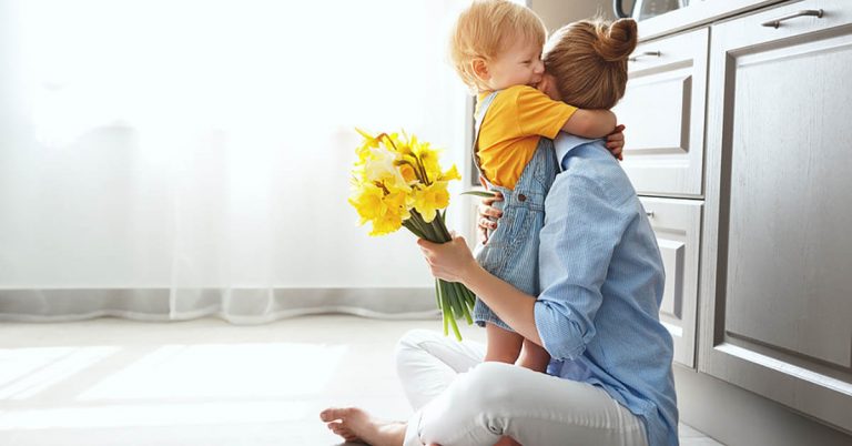 A woman hugs her toddler while sitting on the floor of a bright airy kitchen.